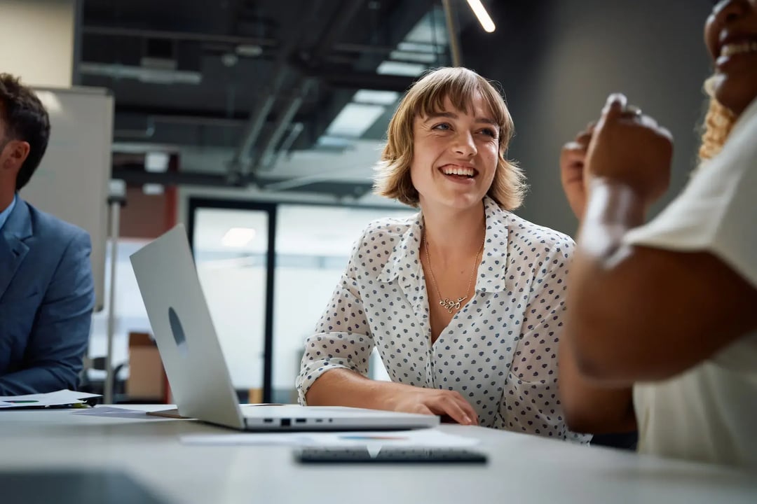 Smiling woman in a modern office environment, engaged in a discussion with colleagues around a table with a laptop